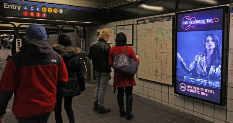 photo of people walking through a subway terminal with an ad displayed on an LCD screen on a wall